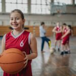 Young girl in a red jersey holds a basketball while wearing braces inside on a basketball court
