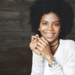 Curly-haired woman wearing braces and a white shirt smiles against a wooden wall